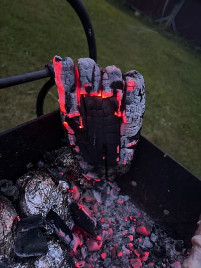The image depicts a close-up view of a fire pit with glowing embers and burning logs. The central subject is a piece of charred wood, emitting a vibrant red glow from within its cracks, indicating intense heat. Surrounding the wood are other pieces of charcoal and some food items wrapped in aluminum foil, suggesting a barbecue setting. The background is softly blurred, revealing a grassy area and what appears to be a fence or structure, possibly indicating an outdoor residential environment. The overall scene is dark, with the light from the fire being the dominant illumination source. The location is described as Манихино Снт, Russia. There are no people visible in the image.