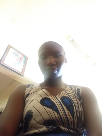 An upward-angled indoor selfie features a person with dark skin and a bald or closely-shaven head, looking directly into the camera. The subject is wearing a sleeveless top with a light-colored base, adorned with a repeating pattern of black lines and distinct blue, oval-shaped designs, reminiscent of African print fabric. Intense overhead lighting creates a bright glare that overexposes parts of the upper background and casts shadows across the subject's face, particularly around the nose and mouth, making their expression appear neutral.
The background reveals a white ceiling with visible grid lines. To the left, a framed picture hangs on a light-colored wall. Although overexposed, the faint image within the frame shows a person's face, possibly wearing headwear, positioned next to vertical stripes of green, white, and red, strongly suggesting the Nigerian flag. To the upper right, a section of an air vent or conditioning unit is visible on the ceiling. The environment appears to be an interior space, likely a building or office, in Nigeria, aligning with the provided location context. No discernible text is visible.