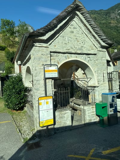 In the mountain village of Lavertezzo, Verzasca, Switzerland, a stone building with a slate roof stands next to a bus stop. The building has arched openings. A bus schedule is posted on a signpost, and a green recycling bin and a ticket machine are visible nearby. The scene is bathed in sunlight, with a clear blue sky above. The surrounding area is mountainous, with trees and vegetation covering the slopes. The ground is paved with asphalt and stone.