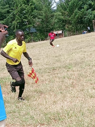 The image depicts an outdoor football (soccer) scene in Lwandeti ward, Kenya. In the central foreground, an adult male identified as an assistant referee runs rightward, wearing a yellow short-sleeved shirt, black shorts, black knee-high socks, and black athletic shoes with white stripes. He holds a red and yellow checkered flag in his right hand. A dark wristband is visible on his left wrist. To the right, a second adult male, a player, wears a red short-sleeved jersey with a white number "10" on the back, dark shorts, and orange athletic shoes. He is in motion near a white football on the field. In the far background, two indistinct figures are visible near a white goal frame, one standing in a white shirt and another seated or crouched. The playing surface consists of dry, light-brown grass with sparse green patches. A dense line of green trees forms the backdrop, partially obscuring a small wooden structure and another building. The top-left corner of the frame shows part of a hand, possibly holding a dark-colored object such as a smartphone.