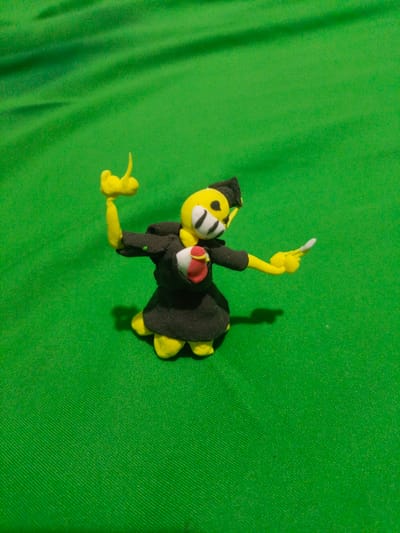 A yellow, clay figurine of a cartoon character with black clothing and a red accent on its chest is positioned on a green, textured surface. The figurine has a black cap and its face features white markings. It is posed with its arms outstretched; one arm is raised and bent, while the other is extended forward, holding a thin white object. The figurine casts a shadow on the green surface. The background is entirely the green textured surface, with subtle diagonal folds. The scene is not located in the city of Gampong Blang, Indonesia.