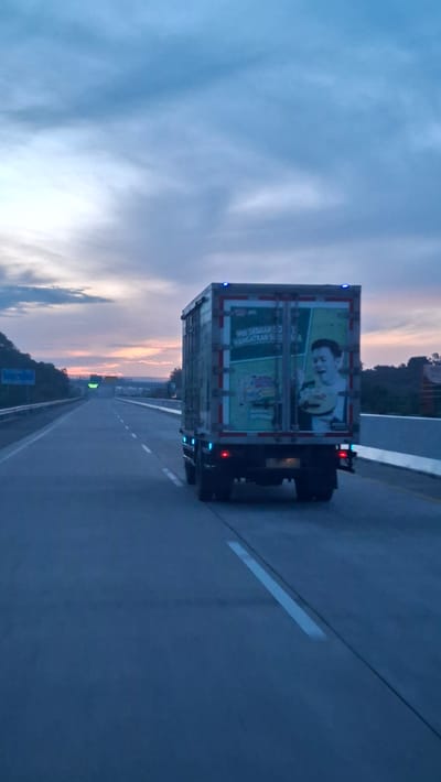 The image shows a truck on a highway at dusk. The truck has an advertisement on its rear depicting a man eating noodles. The sky is cloudy with a hint of orange and pink from the setting sun. The highway is paved and has white lane markings. Trees are visible on the sides of the road. The location is identified as Lambarotunong, Indonesia.