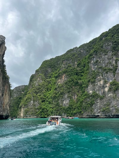 The image depicts a scenic view of limestone cliffs covered in lush green vegetation, jutting out of turquoise-colored water. A white motorboat with several passengers is in the foreground, creating a foamy wake as it moves away from the viewer. In the background, other boats are visible near the shore. The sky is overcast with gray clouds. The location is Haad Yao, Thailand.