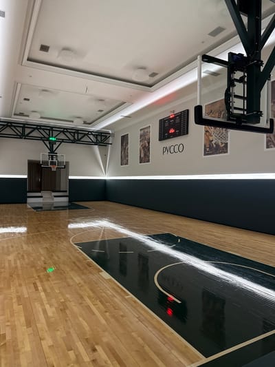 The image shows an indoor basketball court located in Zhukovka, Russia. The court has a polished wooden floor with dark green accents. A basketball hoop with a glass backboard is mounted on a dark green structure. A digital scoreboard displays "17:51" in red digits. The walls are painted dark green, and there is a white illuminated strip running along the top of the walls. Several framed pictures featuring basketball players hang on the wall. The ceiling is white with recessed lighting and ventilation grates. A set of stairs leads to a doorway in the background.