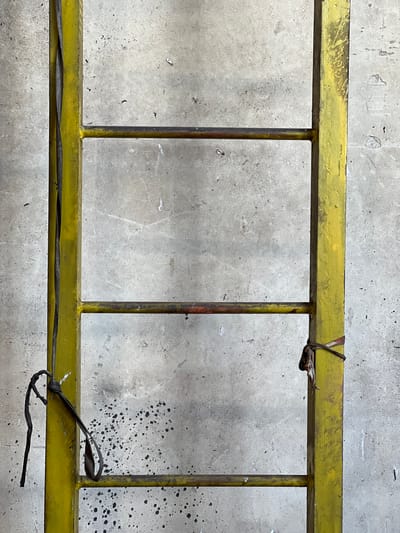 A close-up, vertical shot shows a section of a weathered yellow metal ladder against a textured, light-gray concrete wall. The ladder has three visible horizontal rungs, with the lower two showing some rust. A thick, dark cable is secured to the left vertical support of the ladder with a knot. Another tie, made of what appears to be fabric and wire, is wrapped around the right vertical support near the middle rung. Black speckles are scattered across the concrete wall. The lighting is even, with no distinct shadows. The image appears to be taken outdoors, possibly in an industrial or construction area. The location is identified as Lanžhot, Czechia, but no specific landmarks or features confirm this context within the frame.