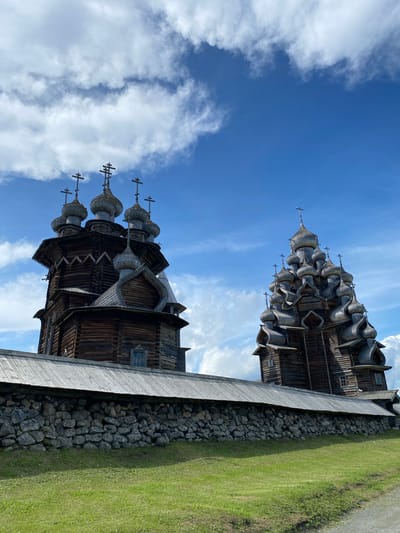The image captures a scene in Boyarshchina, Russia, featuring two historic wooden churches against a bright blue sky with scattered white clouds. The church on the left is a multi-tiered structure topped with seven onion domes, each adorned with a cross. Its dark wooden construction is visible, with intricate details in the rooflines and small windows. A lower wooden roofline extends from this church, supported by a stone wall constructed of irregular grey rocks. The church on the right is even more elaborate, with a complex arrangement of numerous onion domes stacked in a cascading fashion, also topped with crosses. This structure also displays detailed wooden architecture and small windows. In front of the stone wall, a patch of lush green grass slopes gently downwards. The overall impression is one of architectural grandeur set within a natural, open environment. There are no people visible in the image.