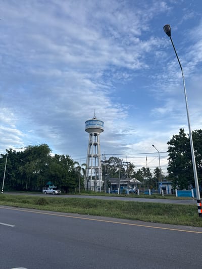 The image depicts a roadside scene in Pak Phraek, Thailand. The primary subjects are a tall water tower and a section of asphalt road. The water tower is white with blue lettering and a conical top, supported by a metal lattice structure. It is positioned behind a line of trees. The road is dark gray with a yellow line marking its edge and a white line indicating a lane. The surrounding environment includes lush green grass and various trees, some of which are palm trees. In the background, there are buildings, power lines, and a street light. A silver pickup truck is parked on the side of the road, partially obscured by foliage. The sky is a prominent feature, filled with scattered white clouds against a bright blue backdrop, suggesting it is daytime, possibly late morning or early afternoon. The overall mood is serene and somewhat rural. The visible text on the water tower, in Thai script, reads "การประปาส่วนภูมิภาค," which translates to "Provincial Waterworks Authority."