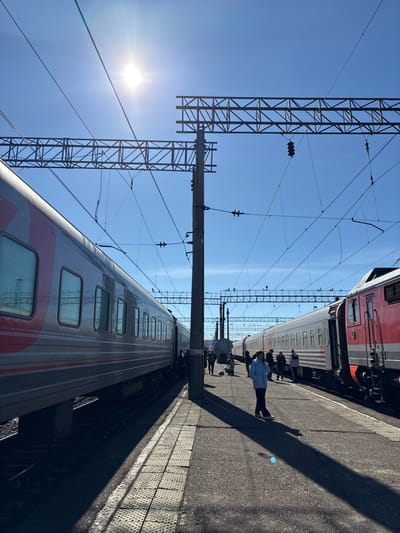 The image depicts a bright, sunny daytime scene at a train station in Kropachyovo, Russia. Two passenger trains are stationary on parallel tracks, separated by a concrete platform. A silver-grey passenger train with a distinctive red stripe runs along the left track, its doors appearing open as several individuals stand nearby, potentially boarding or alighting. On the right track, a red locomotive is coupled to silver-grey passenger cars, with multiple people standing along its side on the platform.
Approximately 10-15 individuals are present on the grey platform. In the immediate foreground, a person wearing a light blue jacket and dark trousers walks towards the lower right, casting a long, distinct shadow. Further down the platform, additional individuals are visible, some with luggage, including a notable red bag near the center.
A tall, dark grey utility pole stands centrally on the platform, supporting an intricate overhead catenary system comprising numerous electrical wires and metal truss structures that extend across the clear blue sky. The sun, a strong light source in the upper left, creates prominent shadows, particularly from the central pole and the foreground individual, stretching across the platform. In the background, additional railway infrastructure, including more overhead gantries and poles, is visible. The platform itself is constructed of concrete, with a textured, lighter-colored tactile strip along its edge.