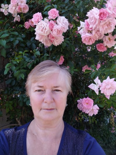 A medium shot shows a woman standing in front of a rose bush in Novoselivka, Ukraine. The woman has short, blonde hair and is wearing a dark blue shirt with lace detailing on the shoulders. She is looking directly at the camera. The rose bush behind her is filled with pink roses in various stages of bloom, and lush green leaves. The background is slightly blurred, suggesting the focus is on the woman and the roses. The overall scene is one of natural beauty and a peaceful outdoor setting.
