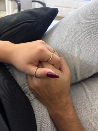 The scene depicts two hands clasped together in an indoor setting, located in the city of Maale Adumim, Palestinian Territory. The hand on top, lighter in skin tone, displays almond-shaped fingernails painted a deep burgundy. This hand wears two rings: a gold-colored band with a twisted, stone-set design on the ring finger, and a thin, stone-set gold-colored band on the pinky finger. The hand beneath, darker in skin tone, exhibits visible hair on its dorsal surface and wrist. Surrounding elements include a black, zippered cushion in the upper left, light gray textured fabric (consistent with denim) on the right side, and dark, ribbed fabric in the lower left. The background is out of focus, indicating an interior location.