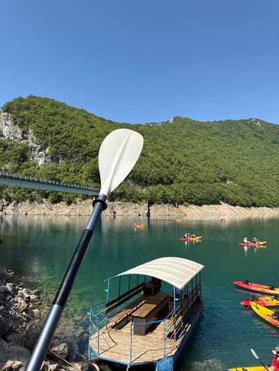 The image depicts a scenic view of the Piva Lake, near the town of Bezuje, Montenegro. In the foreground, a paddle from a kayak is prominently displayed, angled upwards and to the left. Behind the paddle, a clear turquoise lake stretches out, reflecting the bright blue sky and the lush green, tree-covered mountains that flank the water.

Several kayaks, in vibrant red and yellow, are scattered across the lake, with individuals visible in most of them, engaged in kayaking. A small, covered boat with wooden seating and a white canopy is moored near the shore on the right side of the frame. In the background, a large bridge spans the lake, with a railway track running across it. The shores are rocky and lined with sparse vegetation, contrasting with the dense forestation on the mountainsides. The overall scene captures a serene outdoor activity in a breathtaking natural landscape.