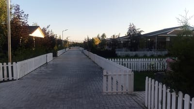 A paved walkway lined with white picket fences stretches into the distance, flanked by houses and trees under a clear sky. The walkway is made of grey rectangular pavers laid in a herringbone pattern. On the left side, a white picket fence follows the walkway, with some dark trees and a building with a triangular roof visible behind it. On the right side, another white picket fence runs parallel to the walkway, with the eaves of low-rise buildings with dark roofs visible behind it. The buildings appear to be modern homes or guest houses, with covered patios and large windows. The environment suggests a suburban or resort setting, possibly in a warm climate given the clear sky and greenery. The scene is devoid of people or any discernable activity. The lighting indicates it is either early morning or late afternoon, with the sun casting a warm glow on the scene. The location is identified as Choktal, Kyrgyzstan.