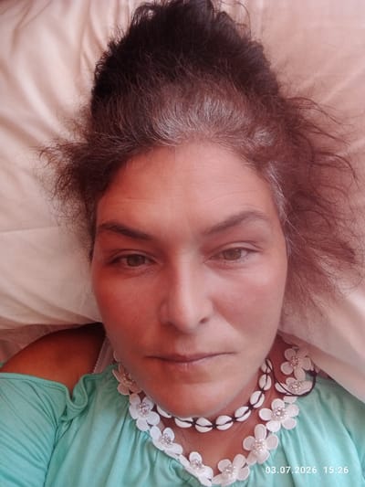 This is a close-up, top-down, selfie-style portrait of a woman lying on a light-colored, wrinkled surface, possibly a bed or couch. The woman has medium-toned skin, with some gray hairs mixed in her dark, somewhat messy hair, which is pulled up. She is wearing a light teal, ruffled off-the-shoulder top and a decorative necklace made of white fabric flowers with small rhinestones and small cowrie shells. The lighting appears warm, possibly from natural light. A timestamp in the lower right corner reads "03.07.2026 15:26". There is no visible indication of the city of Ardea, Italy within the image itself.