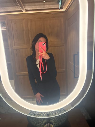 A female individual is depicted in a mirror selfie taken within an indoor facility in St. Christoph, Austria, likely a restroom or changing area. The subject has dark hair and is wearing a black long-sleeved top over a white undershirt, paired with black trousers. Her face is partially obscured by a bright pink smartphone, held in her right hand, which features a matching pink lanyard with silver tips. Her left hand rests on her hip or upper thigh. The mirror itself is U-shaped with integrated white LED lighting illuminating its perimeter. Below the mirror, a dark wooden vanity counter supports a partially visible blue and white patterned ceramic basin with a chrome faucet. The background, reflected in the mirror, consists of light-colored wooden wall paneling and a wooden beamed ceiling. An emergency exit sign is visible in the upper right reflection, alongside a dark rectangular handle on a surface to the right of the subject.