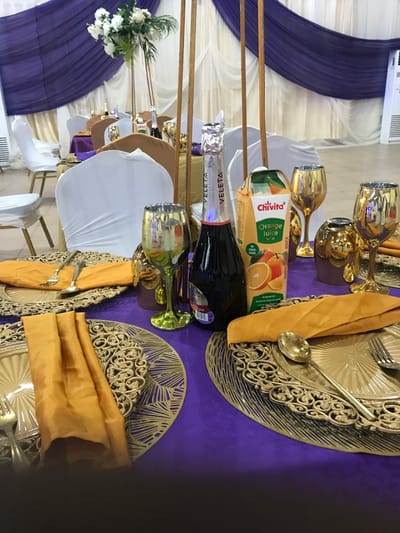 The image depicts a table setting, possibly in a banquet hall, likely located in Ife, Nigeria, given the presence of the "Chivita" brand orange juice, which is common in Nigeria. The table is covered with a purple tablecloth. There are decorative gold charger plates with ornate designs at each place setting. On top of each charger plate is a gold napkin, and utensils (fork and spoon) are placed on the napkin. There are bottles of "VELETA" sparkling wine and cartons of "Chivita" orange juice. There are several gold-colored wine glasses positioned around the bottles and juice carton. In the background, there are white chair covers and purple fabric draping, possibly forming a backdrop or decoration. White flowers are also visible in the background. The focus appears to be on the table setting, suggesting a formal event or celebration.