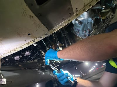 The image shows a person wearing blue gloves performing maintenance on what appears to be the underside of an aircraft. The scene features various mechanical components and structural elements of the aircraft, including metal paneling, riveted joints, and fluid lines. The person is using tools, likely to manipulate or secure parts within the confined space. The lighting suggests an artificial source, possibly from work lights illuminating the area. The background is blurred and indistinct, hinting at a surrounding environment but providing insufficient details to determine whether it is a field or an urban area. It's hard to tell it is in the city of Communauté rurale de Diass, Senegal without further details.