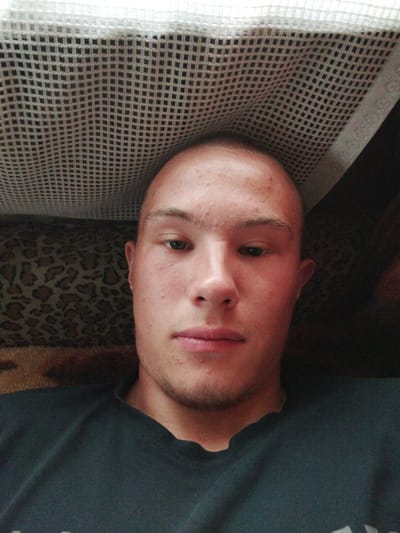 The image is a close-up selfie of a young Caucasian male taken indoors. He has light skin with some acne on his forehead and cheeks, short light brown hair, and a light stubble on his chin. He is wearing a dark t-shirt. The background appears to be a couch with a leopard-print pattern. Above him, a white net-like material is visible, possibly part of a bed or canopy. The lighting suggests an indoor setting. No specific location in the city of Kirensk, Russia, can be identified from the visual information provided in the image.