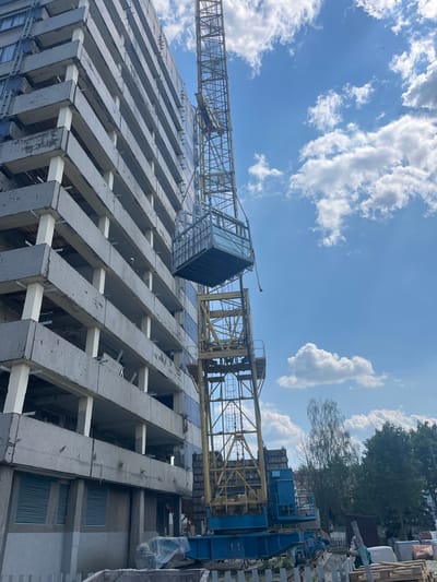 The image depicts a construction site with a tall building under construction and a large yellow crane. The building's floors are visible, characterized by rows of concrete beams supported by lighter-colored beams. A crane with a cabin is positioned over the building, suggesting ongoing construction activities. In the background, there are trees and a blue structure that appears to be part of the construction machinery. The sky is blue with scattered white clouds, indicating daytime and fair weather. The overall scene suggests a busy construction environment. There are no visible text elements in the image.