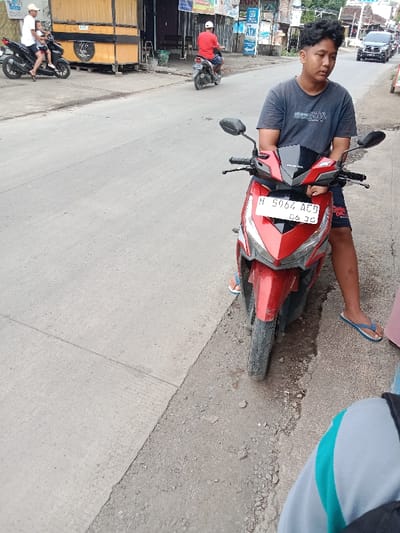 A young man sits on a red Honda motorcycle parked on the side of a road. He wears a dark t-shirt, patterned shorts, and blue flip-flops. The motorcycle has a visible license plate. In the background, another person is riding a motorcycle, and a man in a red shirt rides another further away. A small food stall stands near the roadside. The scene appears to be a casual street scene.