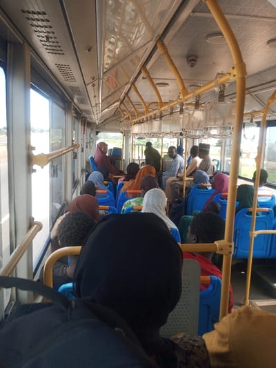The image depicts the interior of a public transportation bus in Zangon Aya, Nigeria, viewed from the rear looking forward. The bus is occupied by approximately 20-25 individuals, predominantly seated.
The interior features rows of blue plastic-backed seats, some with orange or light-colored fabric coverings on the backrests. Above, yellow-painted metal grab rails run horizontally along the ceiling and vertically along the window frames, equipped with clear plastic hanging handholds. The ceiling is light-colored and fitted with circular lighting fixtures, and a security camera is centrally mounted towards the front. Perforated air vents are visible along the upper wall panels. Large windows line both sides of the bus, showing a bright, sunlit exterior with a paved road and green vegetation.
Passengers display a mix of demographics. Many individuals, particularly females, wear head coverings (hijabs or similar) in various colors, including dark maroon, light blue, white, green-patterned, and black. Male passengers are seen wearing shirts; some wear traditional caps. One male in the mid-section wears a light blue long-sleeved shirt. The individual in the immediate foreground, from whose perspective the image is taken, shows the back of a dark-hooded head and part of a black backpack. All observable individuals are either seated or standing passively, indicating routine travel. No active interactions are distinctly visible among the passengers.