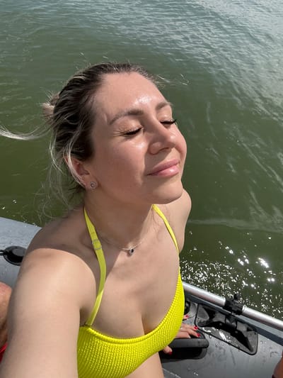 A woman with blonde hair in a bun is wearing a yellow bikini and a necklace. She is on a boat in a body of water. Her eyes are closed, and she is smiling with her face tilted towards the sun. The water is green and appears to be choppy. The boat is gray and has some metal and black plastic components visible.