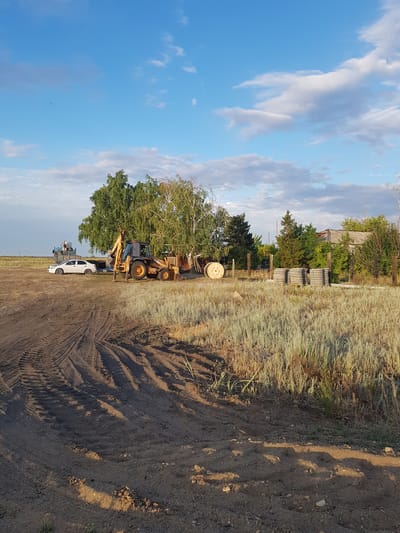 In a rural landscape with a bright blue sky dotted with white clouds, a yellow backhoe loader is parked on a dirt lot. A person is seated in the driver's seat of the backhoe. To the left of the backhoe, a white sedan is parked. Behind the vehicles, a large tree with green leaves stands tall. Further back, several concrete pipes are stacked, and a large wooden spool lies on its side. A rustic fence runs along the edge of the property, with trees and a building visible beyond it. The foreground is dominated by tire tracks imprinted in the dirt. The scene appears to be in the village of Dolen, Kazakhstan.