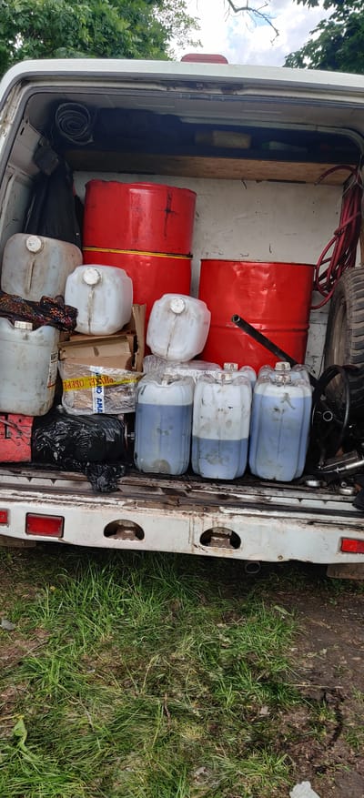 The image shows the open rear of a white van or truck, heavily laden with various items. The interior appears to be filled with cargo, including two red metal barrels, multiple white plastic jerrycans (some with a blue liquid), a spare tire, a steering wheel, a black plastic bag, and cardboard boxes wrapped with tape. The vehicle is parked on grass. The location is in an undefined rural area. There are no people visible in the image.