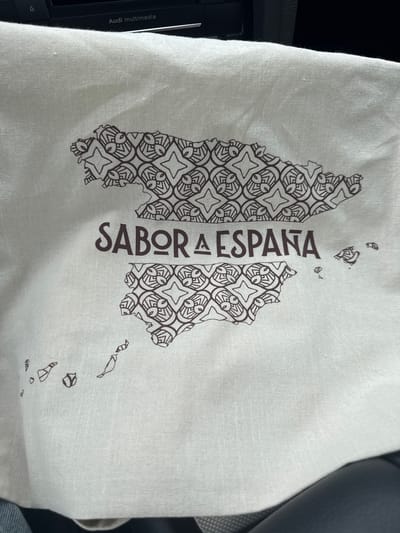 The image shows a close-up view of a white fabric material with a dark brown print. The print depicts a stylized map of Spain, including the Iberian Peninsula and islands. The map is filled with a repeating geometric pattern. Centered below the map of mainland Spain are the words "SABOR A ESPAÑA" in dark brown text. The fabric appears to be a tote bag or similar textile item. In the upper portion of the image, a black car dashboard with "Audi multimedia" visible is blurred. The material is folded and creased, suggesting it is not laid flat. The location in Vellmar, Germany is not depicted or inferable from the image.