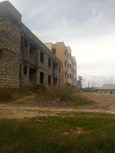 The image depicts an urban development scene in Mtopanga ward, Kenya, under an overcast sky. In the foreground, the ground consists of uneven bare earth interspersed with patches of sparse green grass and dry shrubs.
On the left, a multi-story building constructed from rough-hewn grey concrete blocks is visible, appearing to be in an unfinished state of construction or disrepair. This structure features multiple rectangular openings for windows and doors, lacking frames or glazing, and exhibits exposed rebar elements.
Adjacent to this unfinished structure, on the right, stands a multi-story building painted light yellow. This building appears largely completed, featuring multiple balconies with balustrades and regularly spaced windows.
In the further background, to the right of the yellow building and behind a mound of excavated earth, a low-rise building with a grey roof is partially visible. Sparse vegetation, utility poles, and overhead lines are also present in the distance. No individuals are present in the scene.