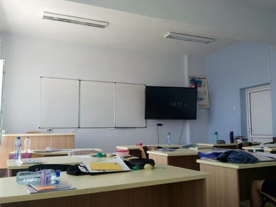 The image depicts the interior of a classroom, likely in Valkosel, Bulgaria, during daylight hours. The room is equipped with multiple wooden desks, each cluttered with various items such as water bottles, notebooks, papers, and stationery. A large whiteboard, divided into three sections, is mounted on the front wall. Adjacent to the whiteboard, a large flat-screen monitor displays the time as 14:01. The walls are painted white and light blue. Two fluorescent light fixtures are installed on the ceiling. A poster with Bulgarian text and imagery, possibly related to national symbols, is displayed on the light blue wall. The scene appears to be unoccupied by students, though a person's legs, wearing dark shorts and a knee, are partially visible in the lower right corner, suggesting an individual is present, possibly a student or teacher. The overall impression is one of a typical school environment, possibly between classes or after instruction has concluded.