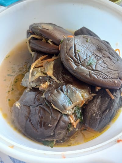 A close-up, overhead shot shows several cooked eggplants submerged in a light-colored broth within a white bowl. The eggplants are dark purple and appear to be stuffed with a filling that includes shredded orange vegetables and green herbs. The broth is slightly oily and has visible specks of herbs throughout. The bowl is white and rests on a surface that is partially visible, appearing to be a white and blue checkered tablecloth. The lighting suggests an outdoor setting during the daytime. The scene is devoid of any people or specific landmarks. The location of Hojamiresen, Turkmenistan is noted in the prompt but there are no visual cues in the image to confirm this location.