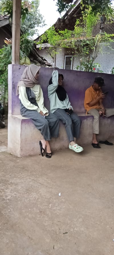 Three people are sitting on a concrete bench in front of a partially-collapsed house, located in Cimaung, Indonesia, at the coordinates -7.074 latitude and 107.547 longitude.  Two women are sitting next to each other, with the woman on the left wearing a light-colored sweater and denim overalls, and the woman on the right wearing a light blue sweater with a black hijab.  The woman on the right is holding a phone in her hand and is looking down at it.  The woman on the left is wearing a light brown hijab and appears to be looking to the right.  The man sitting to the right of the women is wearing a yellow and orange short-sleeve shirt, black pants, and a black baseball cap.  He is looking down at a cell phone he is holding in his right hand.  All three people are wearing sandals.  The house behind the bench is made of concrete with a broken roof.  There is a large tree on the left side of the image. The concrete ground in front of the bench is dirt-colored with some small pieces of trash.