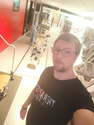 A man with glasses and a goatee is taking a selfie in a gym. He is wearing a black t-shirt with text on it that appears to be backwards and in white and light blue. The text reads "THERAPIST LIFE" with a small heart symbol. He is also wearing a silver chain necklace and a dark bracelet on his left wrist. The background of the gym is slightly blurred due to motion and the camera's focus on the subject. Various gym equipment, including weight machines and what appear to be rowing machines, are visible. The gym has a modern aesthetic with red walls on the left side and bright, possibly fluorescent, lighting. The floor is a light color with darker lines marking out lanes. The overall impression is a well-lit indoor space. The image captures a candid moment, possibly during a workout session.