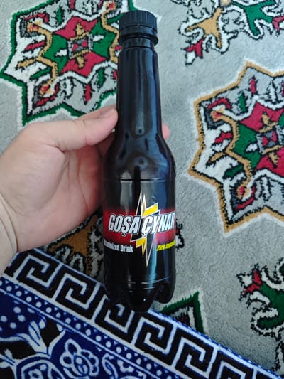 A hand holds a dark-colored plastic bottle of "GOSA CYNAR" flavored drink. The bottle has a red and yellow label featuring a lightning bolt graphic and text in what appears to be Cyrillic and Latin script. The bottle is set against a backdrop of a carpet with a repeating geometric pattern in red, green, yellow, and white. A portion of a blue and white patterned fabric is visible at the bottom of the image. The scene is likely indoors, given the presence of the carpet and the close-up nature of the shot.