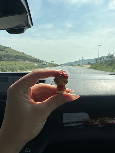A human hand, characterized by extended, manicured fingernails with light polish, occupies the foreground, holding a small tan-colored Hello Kitty figurine between its thumb and forefinger. The figurine is detailed with a red top and a red bow on its head. This is observed from within a vehicle, where a dark dashboard is visible, along with a rearview mirror in the upper left. A compartment on the dashboard contains a white slip of paper with the handwritten numbers "0532" and "376," alongside a partially obscured wrapped chocolate bar. The background, viewed through the vehicle's windshield, shows a multi-lane paved road stretching into the distance under a partly cloudy blue sky. Green, vegetated hills line both sides of the road, with some distant structures on the left hillside and utility poles with overhead lines on the right. Several vehicles are discernible in the distance on the road. The scene depicts a highway or main road environment located within the city of Efeler, Turkey.