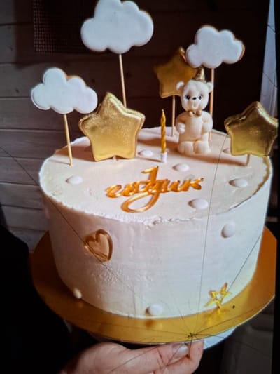 This image shows a white round cake decorated with gold accents, a fondant teddy bear, and gold stars. White cloud-shaped decorations are also attached to skewers, along with the stars and the teddy bear, giving the impression of a sky theme. The cake has gold lettering on its side, which appears to spell out a name, possibly "Luna" or a similar word. The cake sits on a gold cake board. A person's hand is visible on the right side of the frame, holding the edge of the cake board. The background is blurred but appears to be indoors, with a wooden texture suggesting a table or shelf. The lighting is artificial and appears to be coming from above. The overall impression is a celebratory cake, likely for a birthday or baby shower, given the whimsical decorations. No specific time of day or weather conditions are discernible.