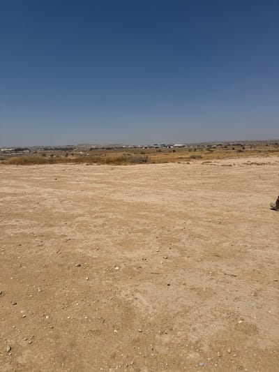 The image displays a broad, dry, sandy landscape with a clear blue sky dominating the left side. The right side of the image features a sandy ground with scattered small rocks and pebbles. In the distance, along the horizon, there is a thin line of vegetation and what appears to be a settlement with some buildings and structures. The lighting suggests it is daytime, likely midday given the brightness and lack of distinct shadows. The scene is arid and open, characteristic of a desert or semi-arid region. There are no people or specific objects clearly visible in the foreground, though a hint of a person's boot is visible in the upper right corner, indicating someone is present or was recently present in the scene. The overall impression is one of a vast, undeveloped natural area adjacent to a populated zone. The location context provided places this scene in the Bnei Shimon Regional Council, Israel, which aligns with the arid landscape depicted.