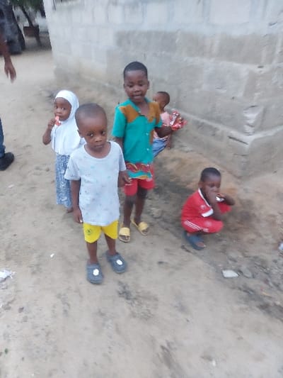 The image depicts an outdoor scene in Mwandege, Tanzania, featuring five young children and a partially visible adult. The setting is an unpaved dirt area situated next to a light grey concrete block wall.
In the foreground, a boy wearing a light-colored t-shirt, yellow shorts, and dark grey closed-toe sandals stands centrally, looking directly towards the camera. To his right and slightly behind him, another boy, dressed in a teal short-sleeved shirt with an orange/brown patterned collar and red patterned shorts with yellow slip-on sandals, stands with his mouth open, looking towards the camera. On the far left, a girl in a white hijab covering her head and torso, paired with a light blue/grey patterned skirt, holds a red and white lollipop stick to her mouth. Behind the second boy, a girl in a light pink top and blue bottoms is partially obscured, holding a package of red items. To the far right, a boy in a red short-sleeved top with white arm stripes and red shorts with white stripes, wearing dark blue slip-on sandals, crouches on the dirt ground with an object near his face. The lower left arm, hand, and a dark blue trouser leg with a black shoe belonging to an adult are visible on the far left of the frame. Small pieces of white litter are scattered on the dirt ground. The scene is illuminated by natural daylight.