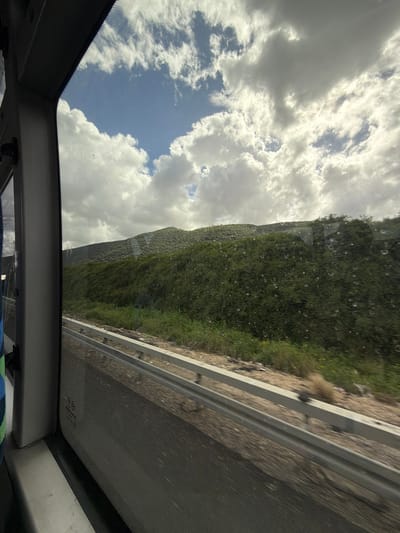The image depicts a view through the open sunroof of a vehicle, likely a car, while traveling on a road. The road, which is bordered by a metal guardrail and a grassy embankment, stretches to the right. The sky, partially covered with clouds and with patches of blue, fills the upper-left portion of the frame. The location is along the road to Zvulun Regional Council, Israel. There are no people visible.