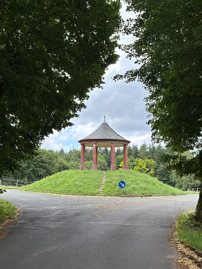 The image shows a circular pavilion with a conical roof, supported by red columns. The pavilion is situated on a grassy hill. A paved road forks in front of the hill, with a blue directional sign indicating a turn to the right. The scene is framed by dense green trees, with a cloudy sky overhead. The location is identified as the Bismarckturm in Heiligenstadt i. OFr., Germany.