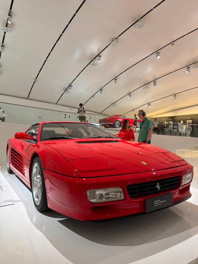 The image depicts the interior of the Museo Enzo Ferrari in Modena, Italy. The foreground is dominated by a bright red Ferrari Testarossa, viewed from a low angle, highlighting its sleek design and iconic details like the side vents and front grille with the Ferrari badge. The car rests on a white, reflective floor that casts subtle shadows. In the middle ground, a couple is observing the Ferrari; the woman wears a red outfit, and the man is in a green shirt and grey shorts, wearing headphones. Behind them, another red Ferrari is displayed, positioned on a raised platform. In the background, several other people are visible, some browsing exhibits, and others appearing to be museum staff. The ceiling features an intricate lighting system with multiple spotlights and curved tracks. The overall environment suggests a modern, well-lit museum exhibit dedicated to Ferrari automobiles.