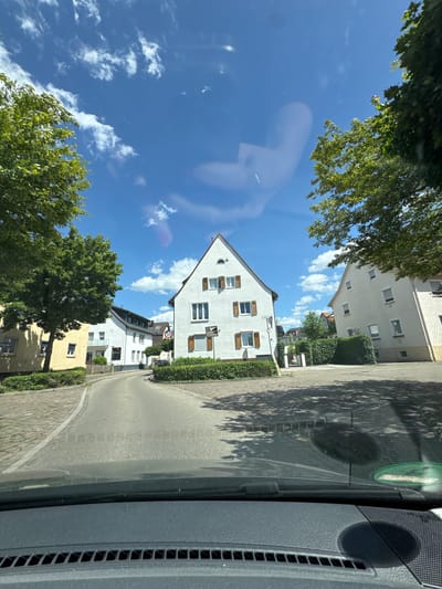 A sunny, blue sky with scattered white clouds is visible above a street in Nürtingen, Germany. The view is from inside a vehicle, looking forward through the windshield. On the left side of the street, a yellow building is partially visible, with trees in front of it. Further down the street, on the right, a white building with brown shutters on its windows is prominently featured. Another white building with a lighter facade stands to the right of the first white building. A 30 km/h speed limit sign is present near the street. The road surface is asphalt, with cobblestone paving on the sidewalks and a portion of the street. Green bushes line the sides of the road in front of the buildings.
