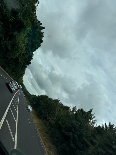 The image depicts a view from a vehicle traveling on a highway in Glenmore, Ireland. The road, marked with white lines and a yellow verge line, curves to the left. Several cars are visible on the road. Dense, dark green trees line both sides of the highway. The sky is overcast with heavy grey clouds. The scene is shot from a low angle, likely from the driver's perspective or a passenger in the front seat.