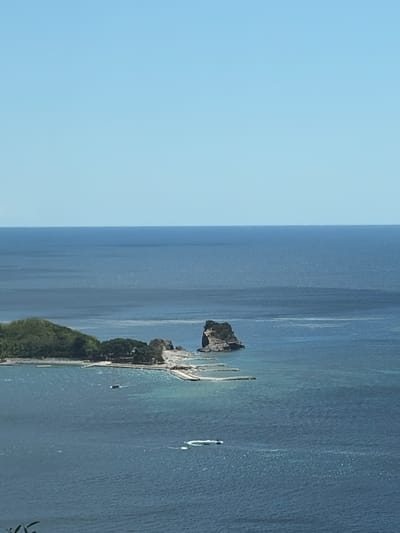 The image shows a panoramic view of a coastal area. In the foreground, a body of deep blue water with some ripples is visible. Several small white objects, likely boats or jet skis, are creating wakes on the water's surface. Further back, a small, lush green island with trees and some structures is situated. Adjacent to the island, a man-made structure, possibly a pier or a breakwater, extends into the water. A prominent rock formation rises out of the sea to the right of the island. The horizon is clearly defined by the meeting of the deep blue sea and a pale blue sky, with no clouds visible. The scene appears to be a scenic coastal landscape, likely in a Mediterranean or similar climate, possibly depicting the area around Košljun, Montenegro, given the description of a small island with a monastery.