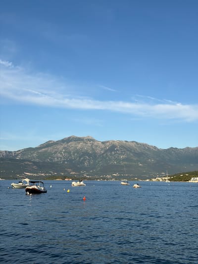 The image depicts a coastal scene, likely a part of Krašići, Montenegro. The view features a mountain range running parallel to the coastline. Clear blue sky is partially covered with long, thin clouds. The sea is calm with visible ripples and the color is a dark blue hue. A number of boats are anchored in the water, appearing along the coastline.