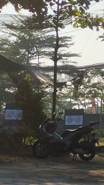 The image depicts an outdoor archery range located in Trosobo, Indonesia. Two archery targets are visible, mounted on dark rectangular backdrops. One target is positioned on the left side of the frame, and another is on the right. Between the targets, a dark-colored motorcycle is parked on a dirt or gravel surface. The background is characterized by trees, some of which are partially obscured by a netted canopy overhead. The overall lighting suggests it is daytime, with a bright sky visible through the foliage.