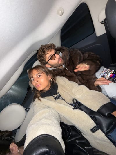 The image, taken in Villeneuve-Loubet, France, features a man and a woman inside a car, captured from a tilted perspective looking upwards. The car's light-colored ceiling and a dark sunroof are visible in the background.
On the left, a man with curly brown hair and a beard is seen wearing dark sunglasses with light frames and a brown, textured, possibly faux fur, hooded jacket. His expression is neutral. Above him, a hand holds a smartphone, which displays a grid or collage of multiple small images on its screen.
Beside the man, a woman with straight brown hair looks towards the camera. She wears a black top or hoodie underneath a cream or beige shearling-lined jacket. A black stylized logo, resembling an 'LV' or 'V', is prominently visible on the front of her jacket. She has an earring in her right ear and her expression is somewhat serious or contemplative. A black bag or strap is draped over her shoulder. Part of another person's head and hair can be seen in the bottom right corner of the frame. The subdued lighting suggests an indoor setting, possibly evening or night.