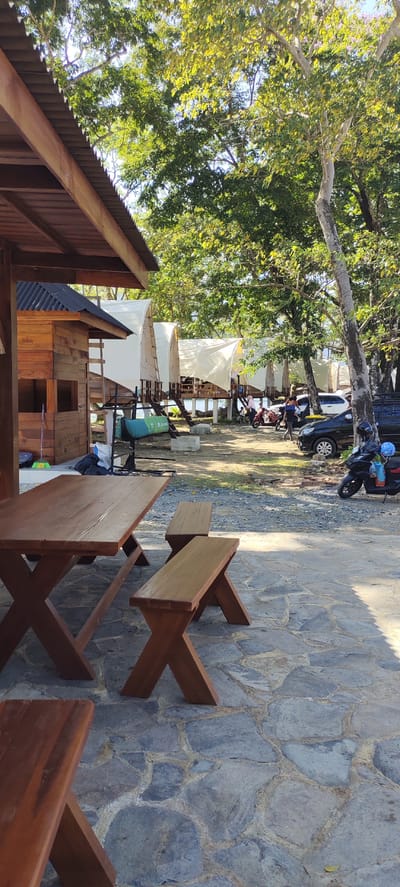 In Lamseunia, Indonesia, a scene unfolds at an outdoor establishment featuring a picnic table and benches in the foreground. Beyond this seating area, a row of canvas-covered tent structures on stilts lines the waterfront. Trees with lush green foliage dominate the background, casting dappled shadows on the ground. To the right, several parked vehicles, including a black car and a scooter, are visible. A person is seen on the scooter, and another individual is walking in the vicinity of the vehicles. A broom leans against a wooden structure on the left side of the image. The ground is covered with a mixture of gravel and flagstone.
