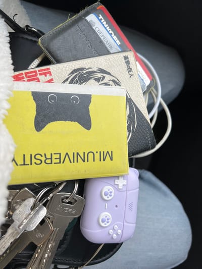 A close-up, overhead view displays an assortment of personal items resting on a lap in the city of Lipitsy, Russia. The scene is static, with no visible individuals engaged in actions.
Forensically, the following objects are identified:
* **Top Left:** A section of white, textured fabric, consistent with a garment or bag lining.
* **Center Foreground:** A yellow rectangular card or item featuring an upside-down black cat silhouette with white eyes. Below this, also upside down, are the black letters "M.L.UNIVERSITY".
* **Below and Behind the Yellow Item:** A dark-colored, possibly leather, wallet or card holder is partially visible. It contains multiple cards.
* A red card with white lettering reads "TINKOFF".
* A light blue card with white lettering reads "СБЕРБАНК".
* Additional obscured cards are present.
* **Behind the Wallet:** A light-colored item, possibly a passport cover or another wallet, displays a black graphic of a stylized head or character, and Japanese text which translates to "Attack on Titan" ("進撃の巨人"). The name "LEVI ACKERMAN" is also partially visible.
* **Entangled:** A bundle of white cables (likely USB or charging cables) is visible behind these items.
* **Bottom Left:** A cluster of metal keys on a key ring. One key prominently displays the brand "AVERE".
* **Bottom Right:** A light purple silicone case, designed to resemble a retro video game controller, is attached to the key ring. It features white control buttons, including a directional pad, action buttons labeled 'A' and 'B', and two analog sticks with white paw print designs.
* **Underlying Surface:** The items are resting on light blue denim fabric, indicating the presence of a person's leg or lap, and a darker, indistinct surface on the right.