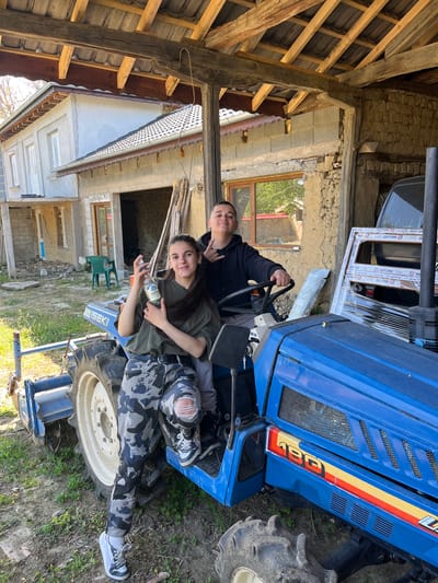 Two people are on a blue ISEKI 180 tractor. One is seated in the driver's seat while the other is reclining on the hood. They appear to be posing for a photo. The woman lying on the hood is holding an unlabeled bottle. The background includes a partially renovated building, a wooden support structure, and various construction materials. The scene is outdoors in what appears to be a rural setting. The location is Rish, Bulgaria.